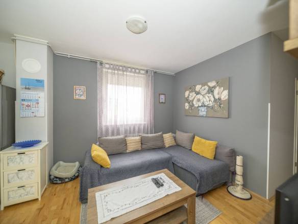 Ugodan sivi dnevni boravak s L-oblikovana sofom, jastucima boje senfa i drvenim stolićem za kavu ispred; prozirne zavjese preko prozora na sivim zidovima.