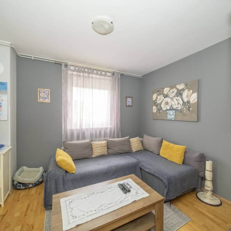 Ugodan sivi dnevni boravak s L-oblikovana sofom, jastucima boje senfa i drvenim stolićem za kavu ispred; prozirne zavjese preko prozora na sivim zidovima.