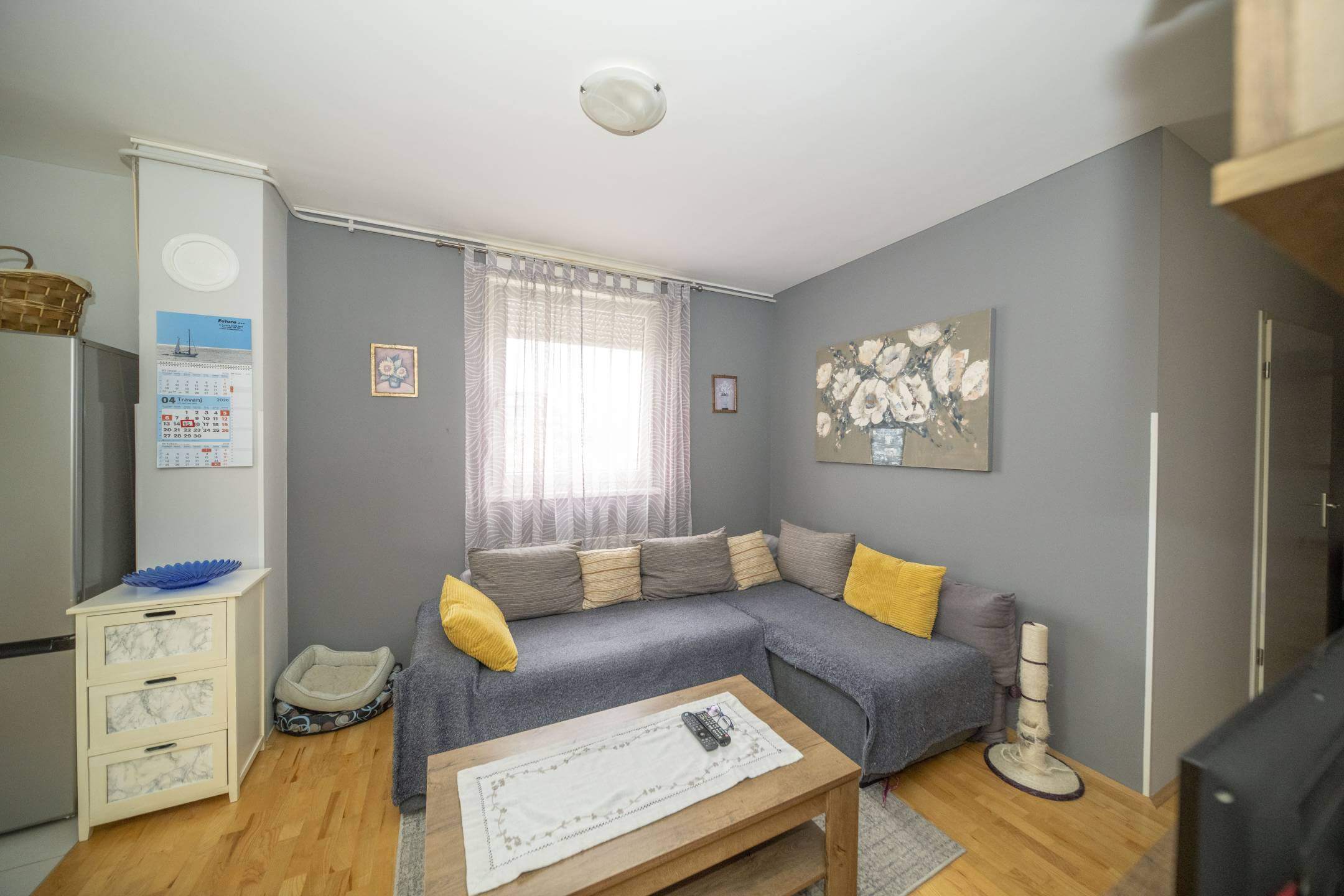 Ugodan sivi dnevni boravak s L-oblikovana sofom, jastucima boje senfa i drvenim stolićem za kavu ispred; prozirne zavjese preko prozora na sivim zidovima.