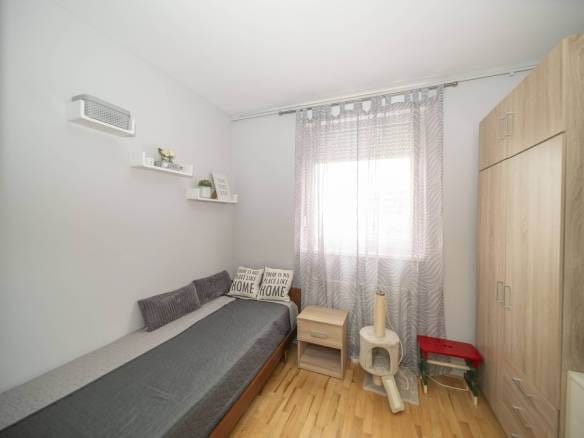 Mala spavaća soba s jednokrevetnim krevetom, sivim poplunom i svijetlim drvenim namještajem; prozor s prozirnim zavjesama s uzorkom ispunjava sobu mekim svjetlom.