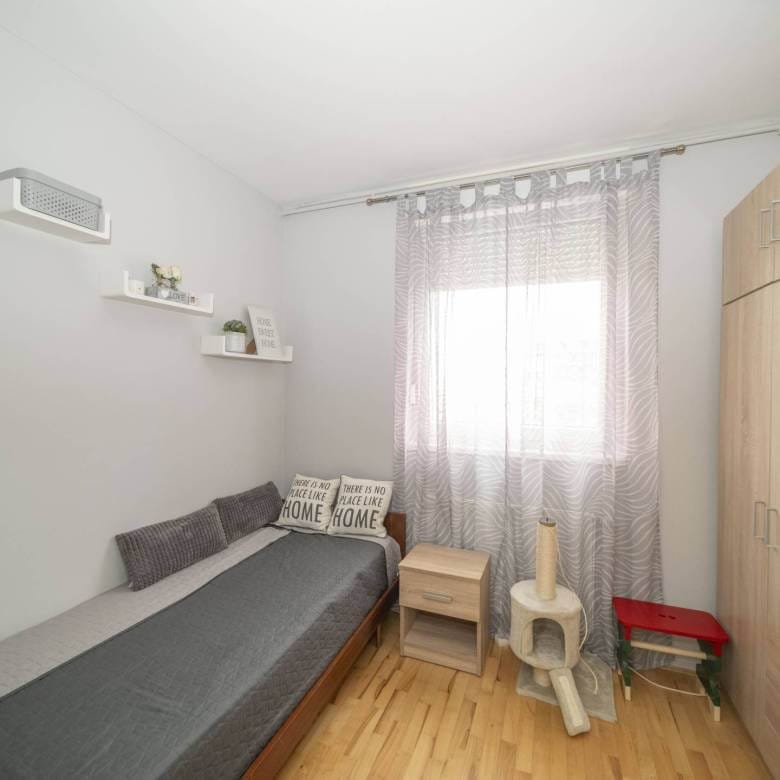 Mala spavaća soba s jednokrevetnim krevetom, sivim poplunom i svijetlim drvenim namještajem; prozor s prozirnim zavjesama s uzorkom ispunjava sobu mekim svjetlom.