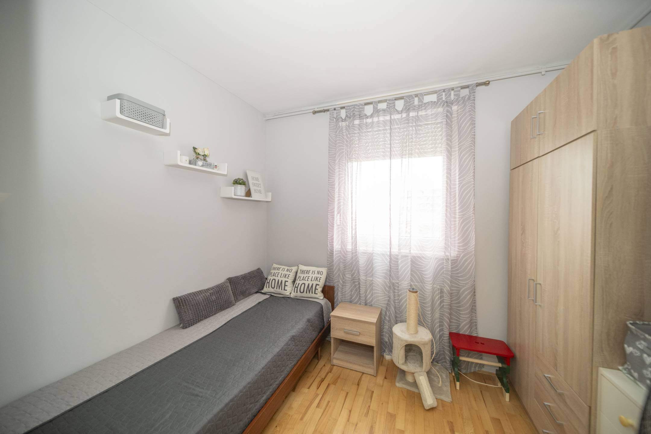 Mala spavaća soba s jednokrevetnim krevetom, sivim poplunom i svijetlim drvenim namještajem; prozor s prozirnim zavjesama s uzorkom ispunjava sobu mekim svjetlom.