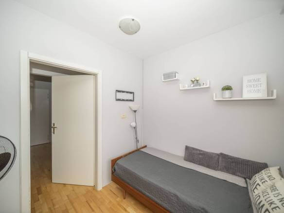 Mala spavaća soba s jednim drvenim krevetom, sivom posteljinom i dva siva jastuka uz svijetlosive zidove, otvorena vrata otkrivaju hodnik.