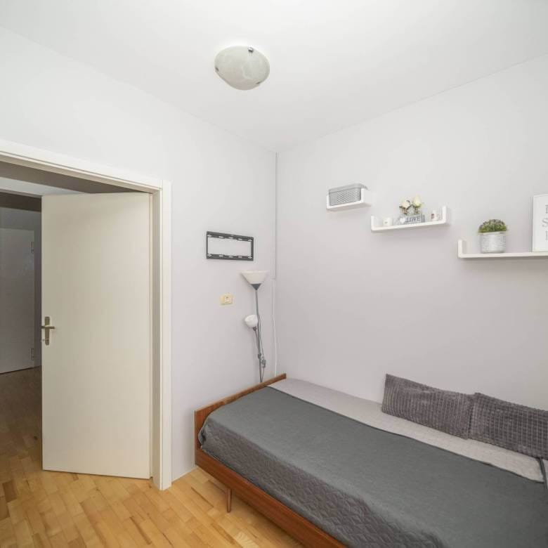 Mala spavaća soba s jednim drvenim krevetom, sivom posteljinom i dva siva jastuka uz svijetlosive zidove, otvorena vrata otkrivaju hodnik.