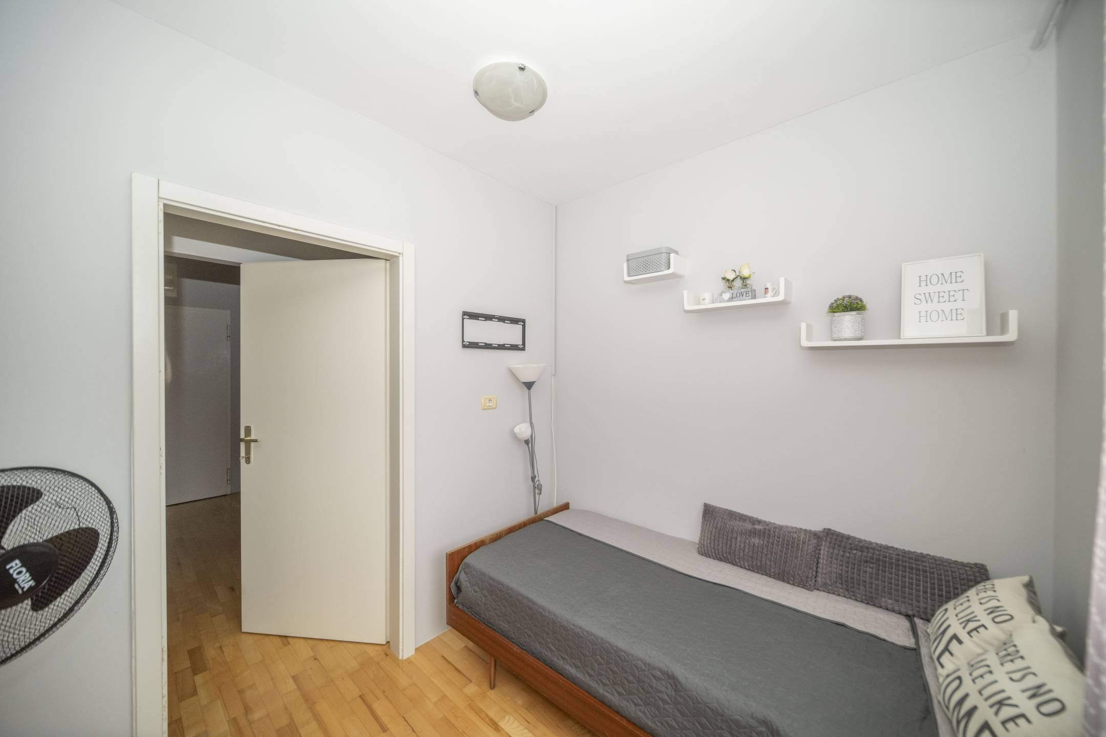 Mala spavaća soba s jednim drvenim krevetom, sivom posteljinom i dva siva jastuka uz svijetlosive zidove, otvorena vrata otkrivaju hodnik.