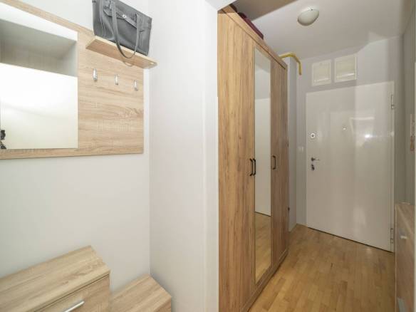 Ulazni prostor sa svijetlim drvenim namještajem, zidnim ogledalom, kukama i visokim ormarom uz bijele zidove