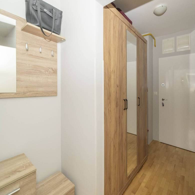 Ulazni prostor sa svijetlim drvenim namještajem, zidnim ogledalom, kukama i visokim ormarom uz bijele zidove