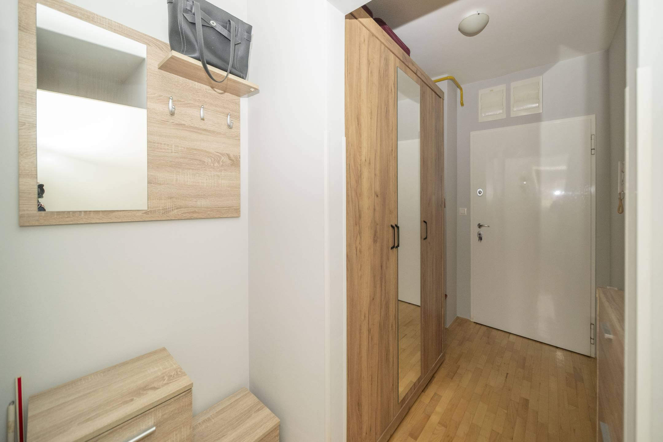 Ulazni prostor sa svijetlim drvenim namještajem, zidnim ogledalom, kukama i visokim ormarom uz bijele zidove