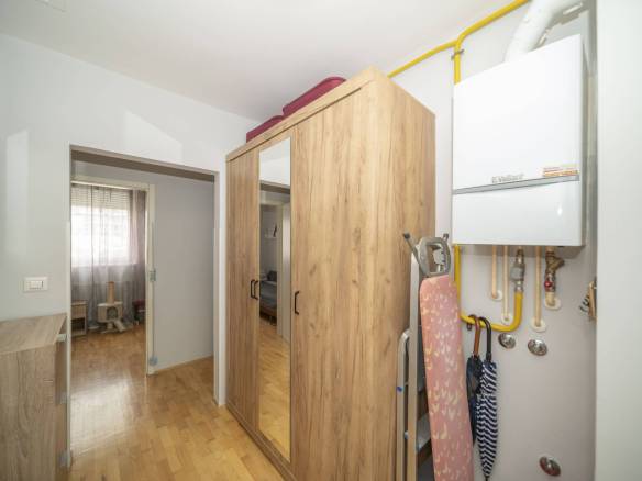 Pomoćni/skladišni prostor s visokim drvenim ormarom, vratima sa zrcalima i daskom za glačanje pokraj bojlera na zidu; žute plinske cijevi prolaze iznad.