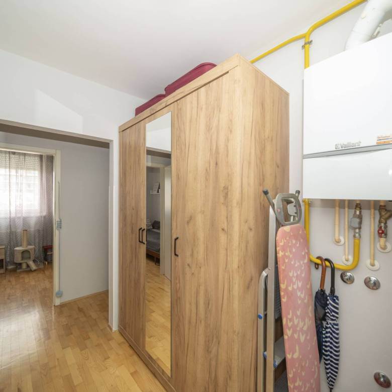 Pomoćni/skladišni prostor s visokim drvenim ormarom, vratima sa zrcalima i daskom za glačanje pokraj bojlera na zidu; žute plinske cijevi prolaze iznad.