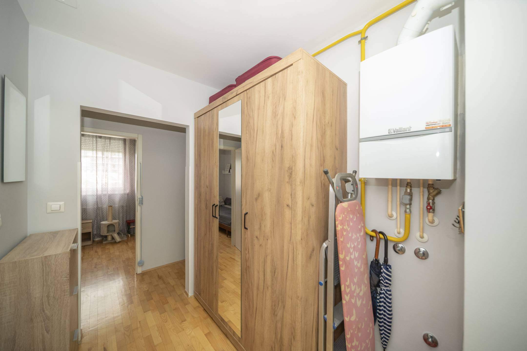 Pomoćni/skladišni prostor s visokim drvenim ormarom, vratima sa zrcalima i daskom za glačanje pokraj bojlera na zidu; žute plinske cijevi prolaze iznad.