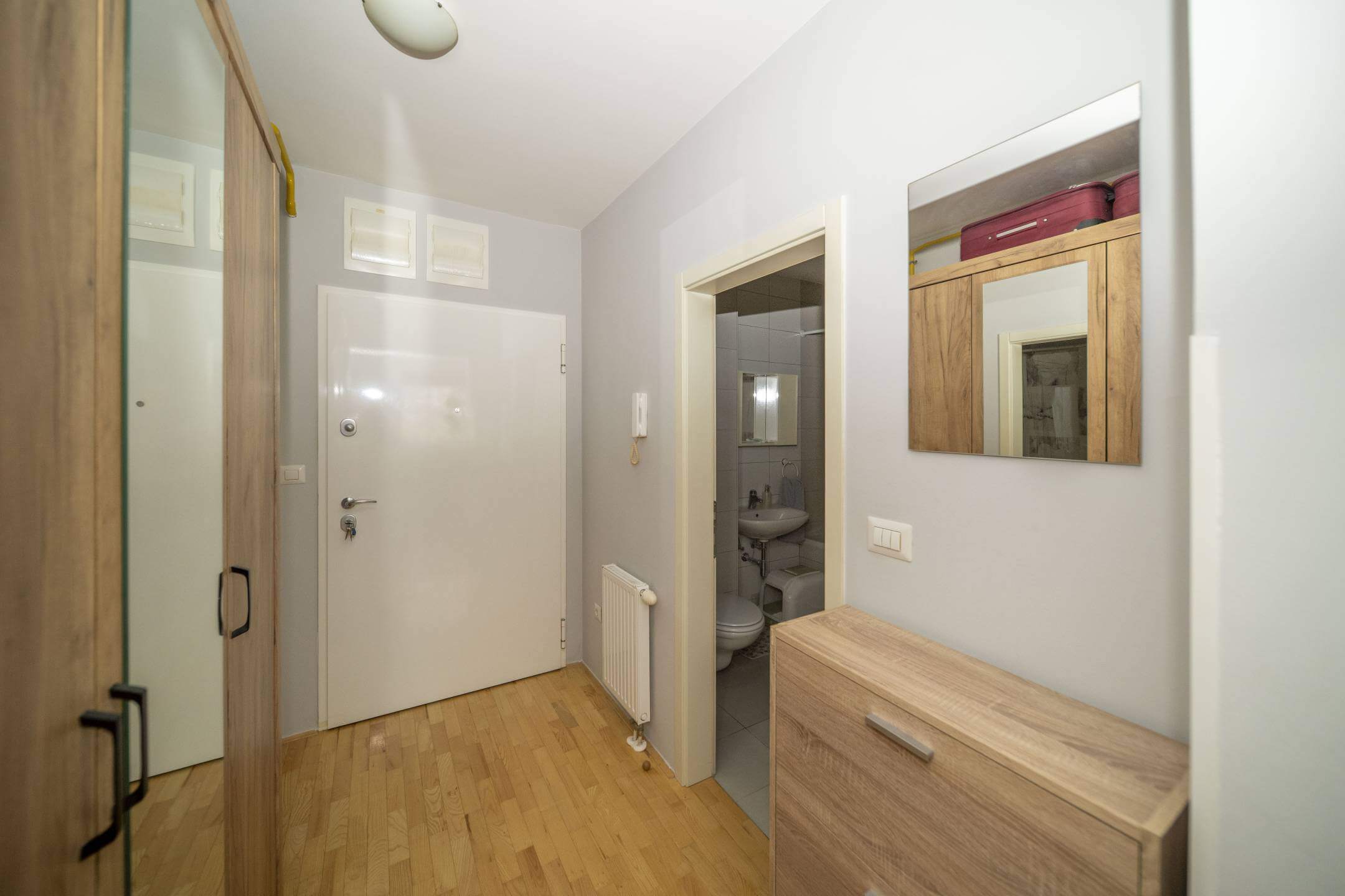 Ulaz u hodnik s bijelim ulaznim vratima, svijetlim drvenim podom, radijatorom i drvenom komodom; vrata kupaonice s desne strane.