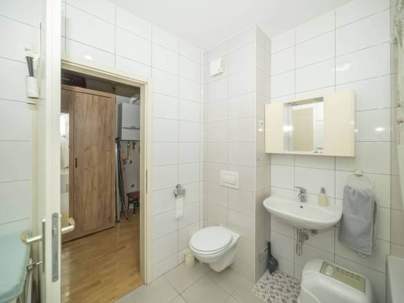 Kupaonica s bijelim pločicama, zidnim umivaonikom i ormarićem s ogledalom iznad, zidnim WC-om, vješalom za ručnike sa sivim ručnikom i vratima koja vode u susjednu sobu s drvenim ormarom u pogledu.