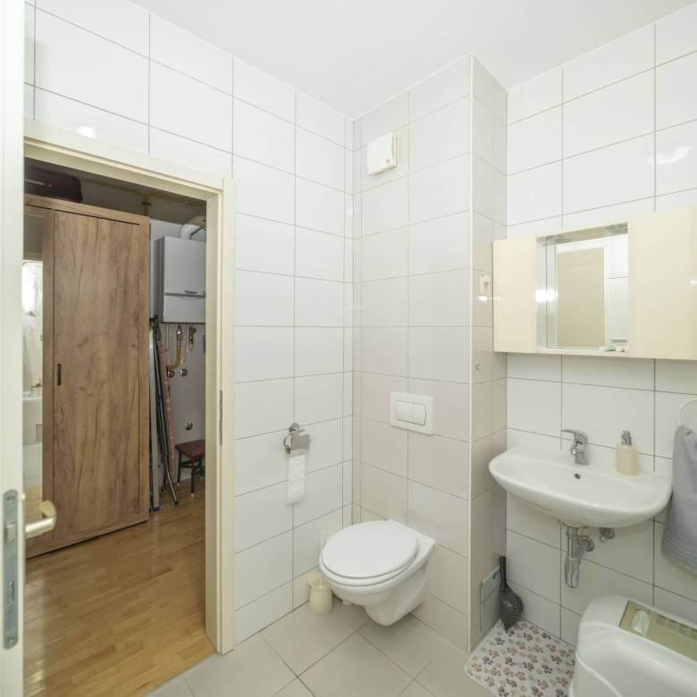 Kupaonica s bijelim pločicama, zidnim umivaonikom i ormarićem s ogledalom iznad, zidnim WC-om, vješalom za ručnike sa sivim ručnikom i vratima koja vode u susjednu sobu s drvenim ormarom u pogledu.