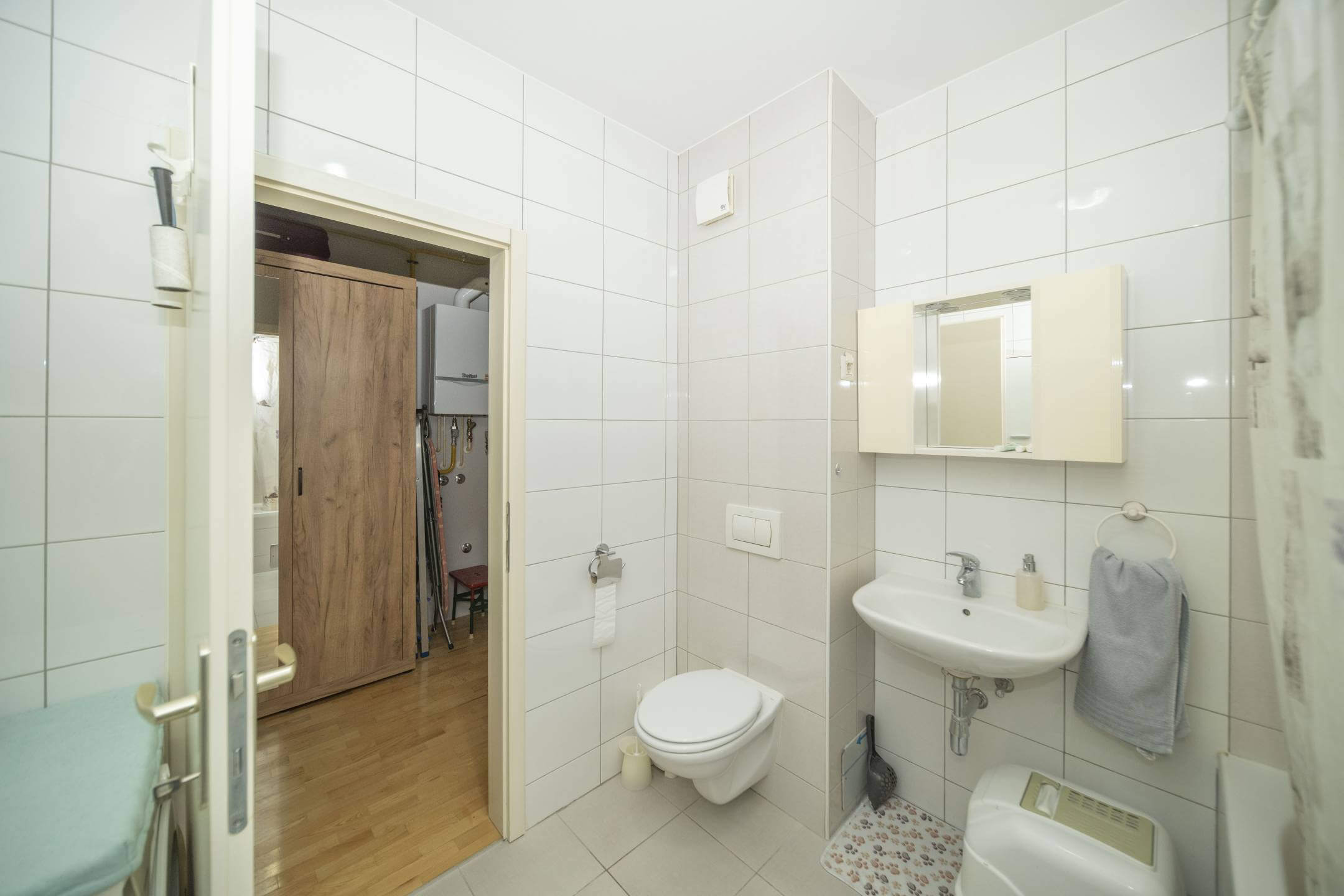 Kupaonica s bijelim pločicama, zidnim umivaonikom i ormarićem s ogledalom iznad, zidnim WC-om, vješalom za ručnike sa sivim ručnikom i vratima koja vode u susjednu sobu s drvenim ormarom u pogledu.