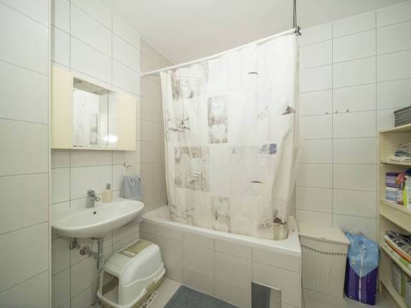 Mala kupaonica s pločicama, bijelom kadom i zavjesom za tuš s uzorkom, umivaonikom, ormarićem s ogledalom i otvorenom policom s ručnicima i priborom.