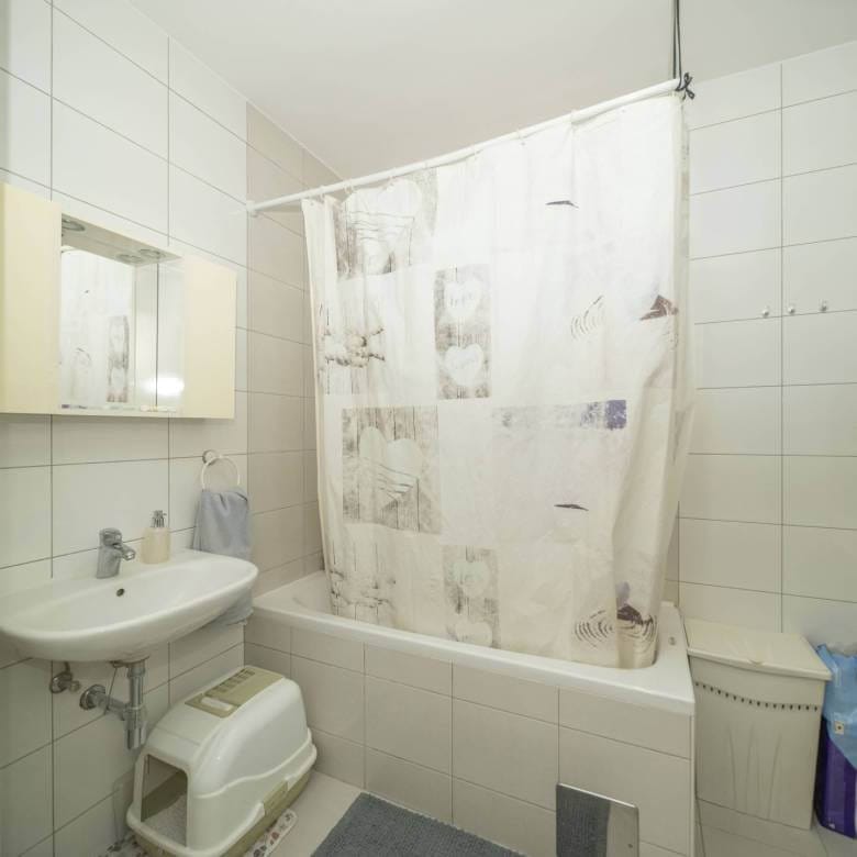 Mala kupaonica s pločicama, bijelom kadom i zavjesom za tuš s uzorkom, umivaonikom, ormarićem s ogledalom i otvorenom policom s ručnicima i priborom.