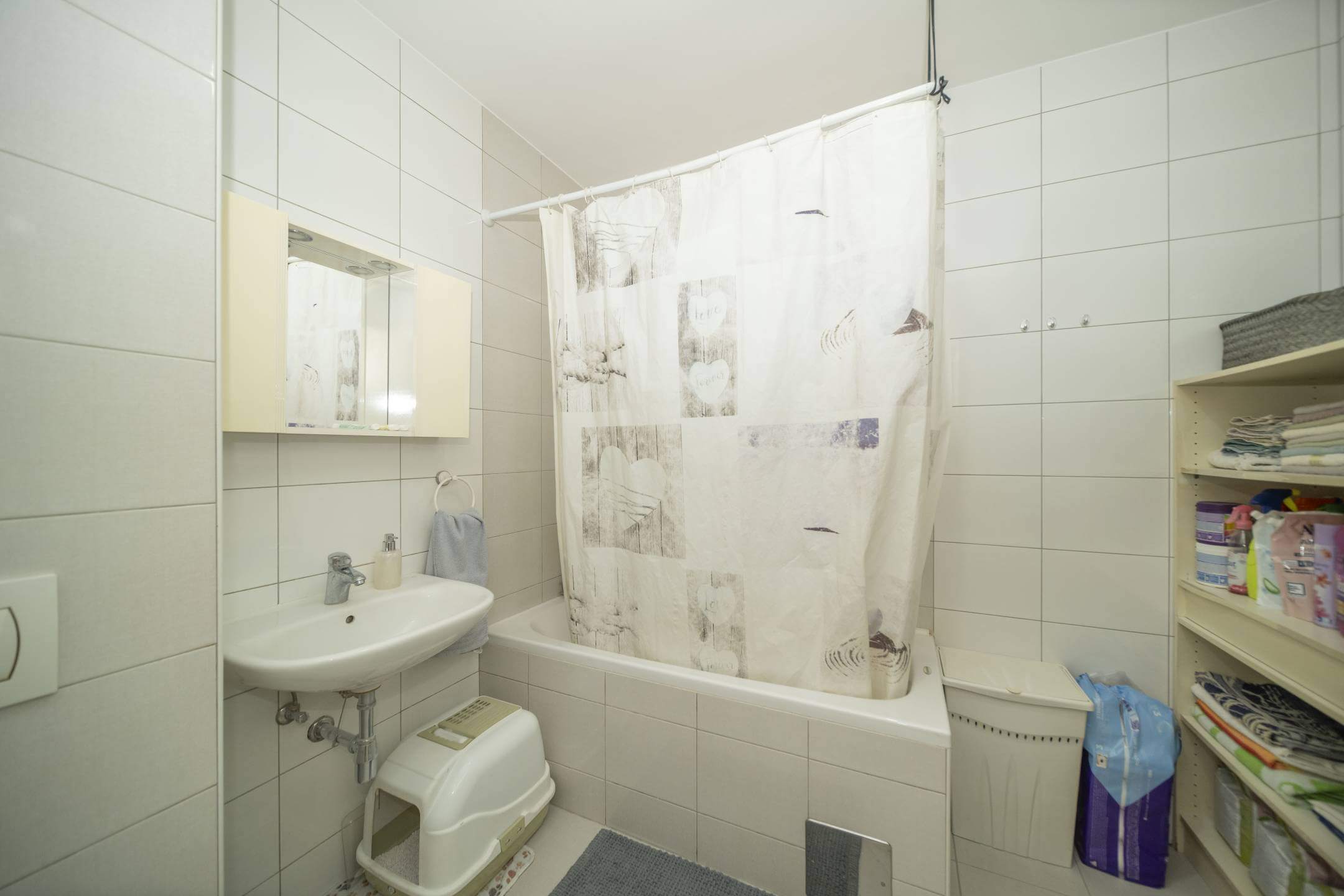 Mala kupaonica s pločicama, bijelom kadom i zavjesom za tuš s uzorkom, umivaonikom, ormarićem s ogledalom i otvorenom policom s ručnicima i priborom.