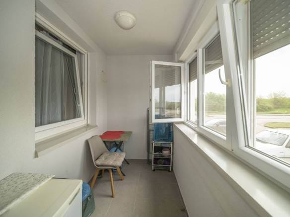 Uski zatvoreni balkon s malim stolom i stolicom, kolicima za odlaganje i velikim prozorima s pogledom na parkiralište.