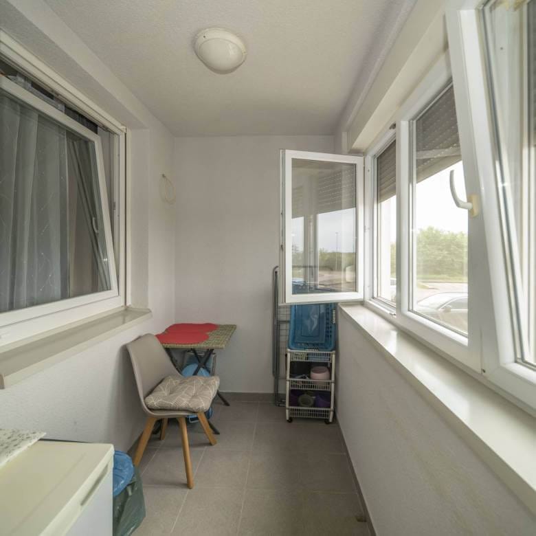 Uski zatvoreni balkon s malim stolom i stolicom, kolicima za odlaganje i velikim prozorima s pogledom na parkiralište.