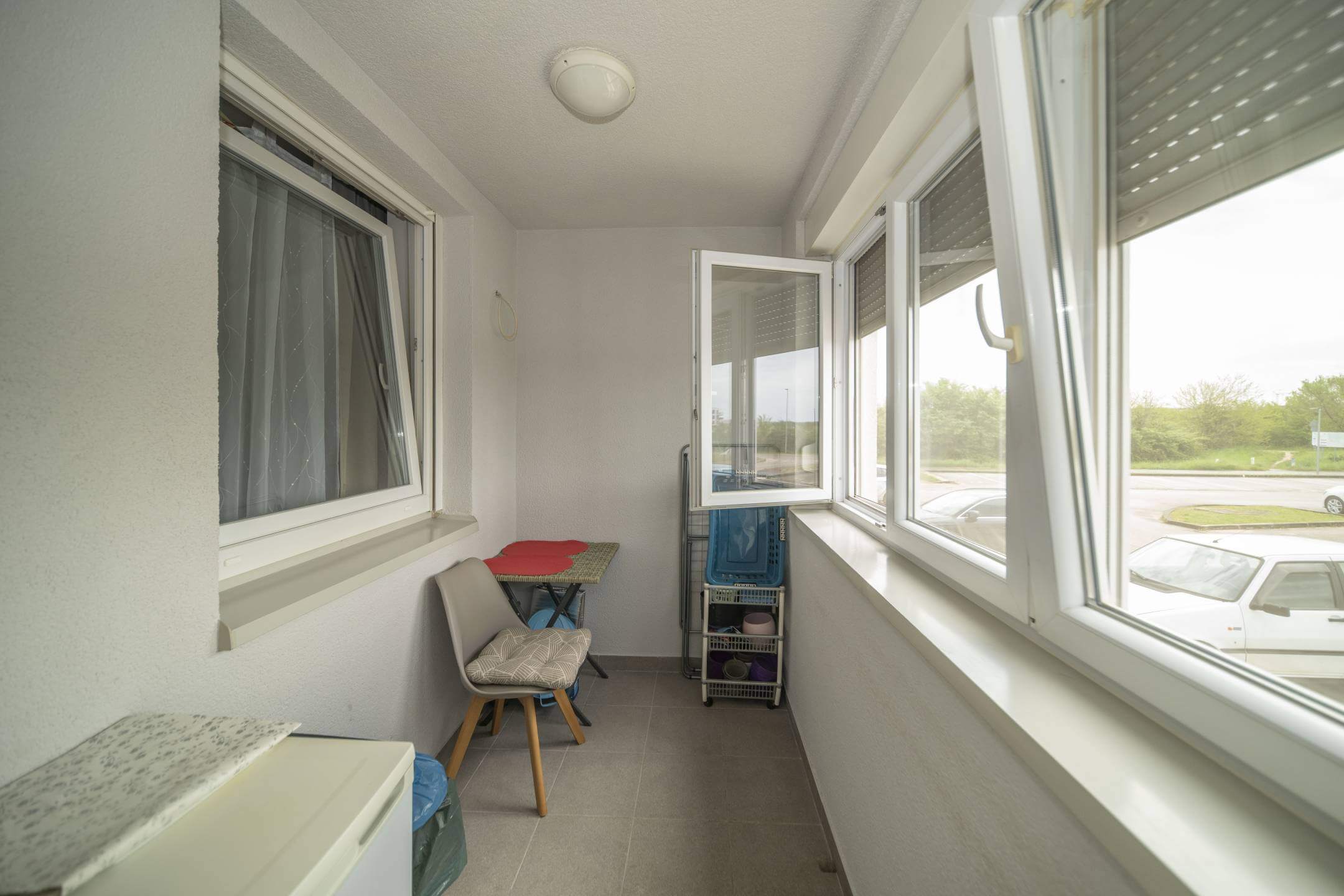 Uski zatvoreni balkon s malim stolom i stolicom, kolicima za odlaganje i velikim prozorima s pogledom na parkiralište.