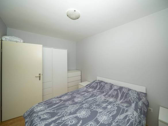 Mala, minimalistička spavaća soba s bijelim okvirom kreveta, sivim cvjetnim prekrivačem i bijelim ormarima uz zid.