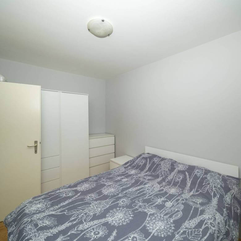 Mala, minimalistička spavaća soba s bijelim okvirom kreveta, sivim cvjetnim prekrivačem i bijelim ormarima uz zid.