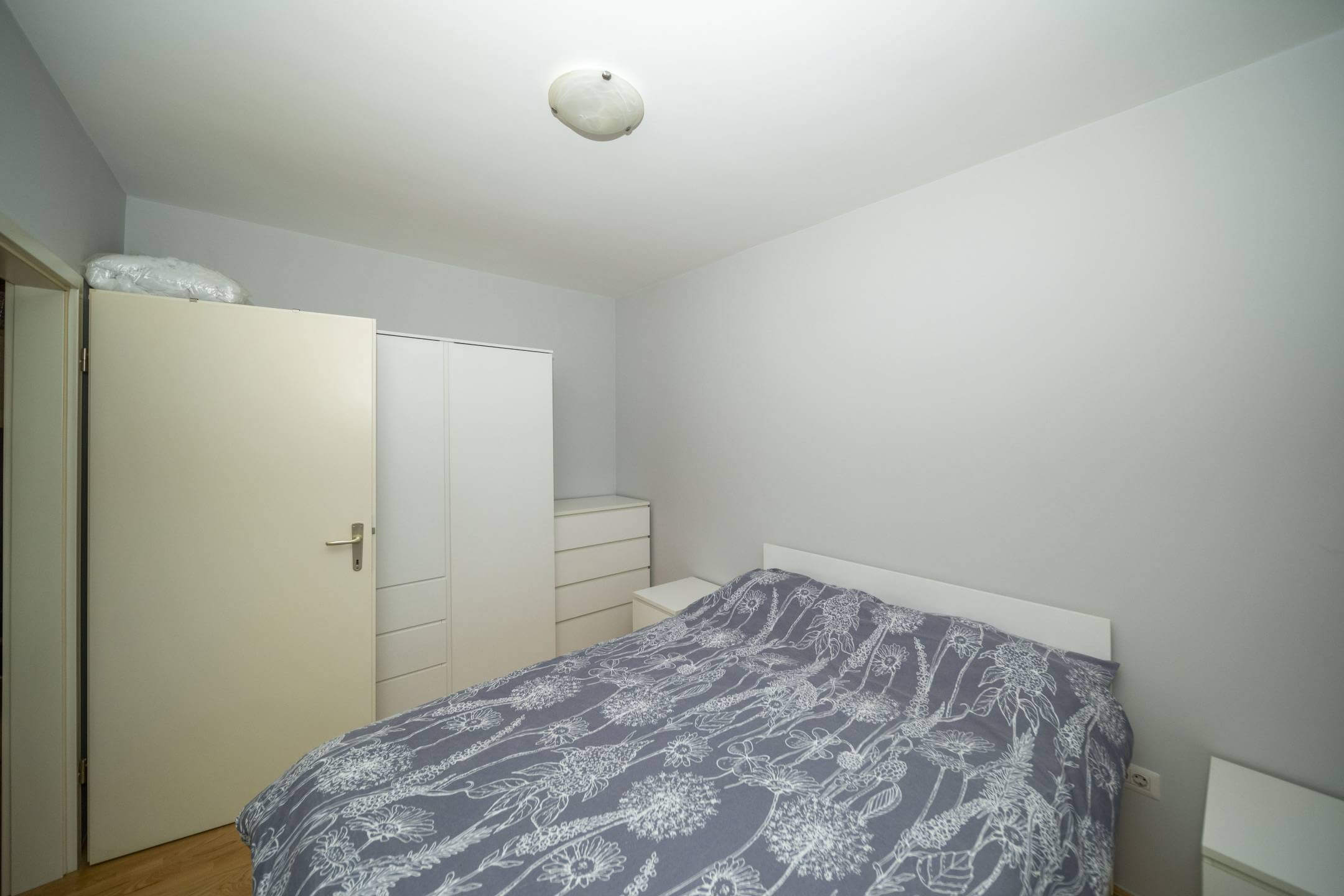 Mala, minimalistička spavaća soba s bijelim okvirom kreveta, sivim cvjetnim prekrivačem i bijelim ormarima uz zid.
