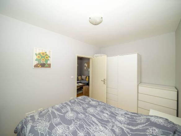 Mala uredna spavaća soba sa sivim cvjetnim prekrivačem, bijelim ormarom i komodom, otvorena vrata otkrivaju dnevni boravak.