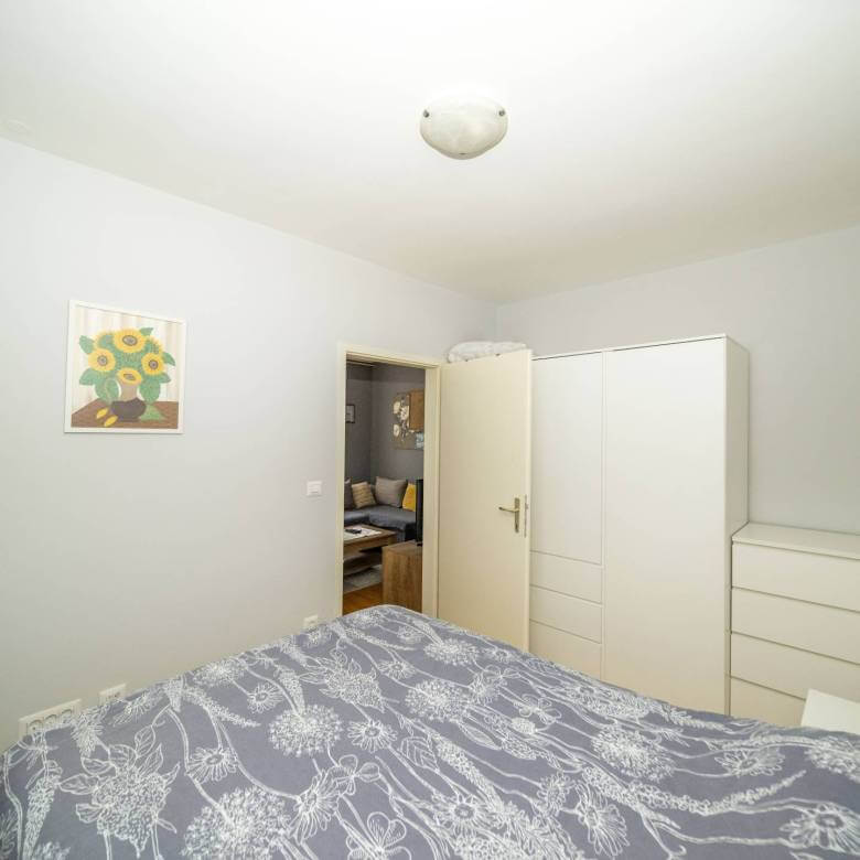 Mala uredna spavaća soba sa sivim cvjetnim prekrivačem, bijelim ormarom i komodom, otvorena vrata otkrivaju dnevni boravak.
