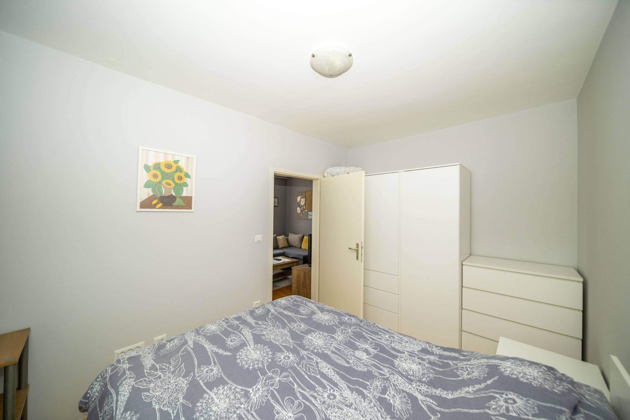Mala uredna spavaća soba sa sivim cvjetnim prekrivačem, bijelim ormarom i komodom, otvorena vrata otkrivaju dnevni boravak.
