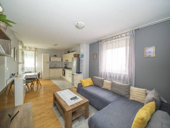 Otvoreni dnevni boravak sa sivom sofom u obliku slova L, žutim jastucima i drvenim stolićem za kavu; kuhinja u pozadini.