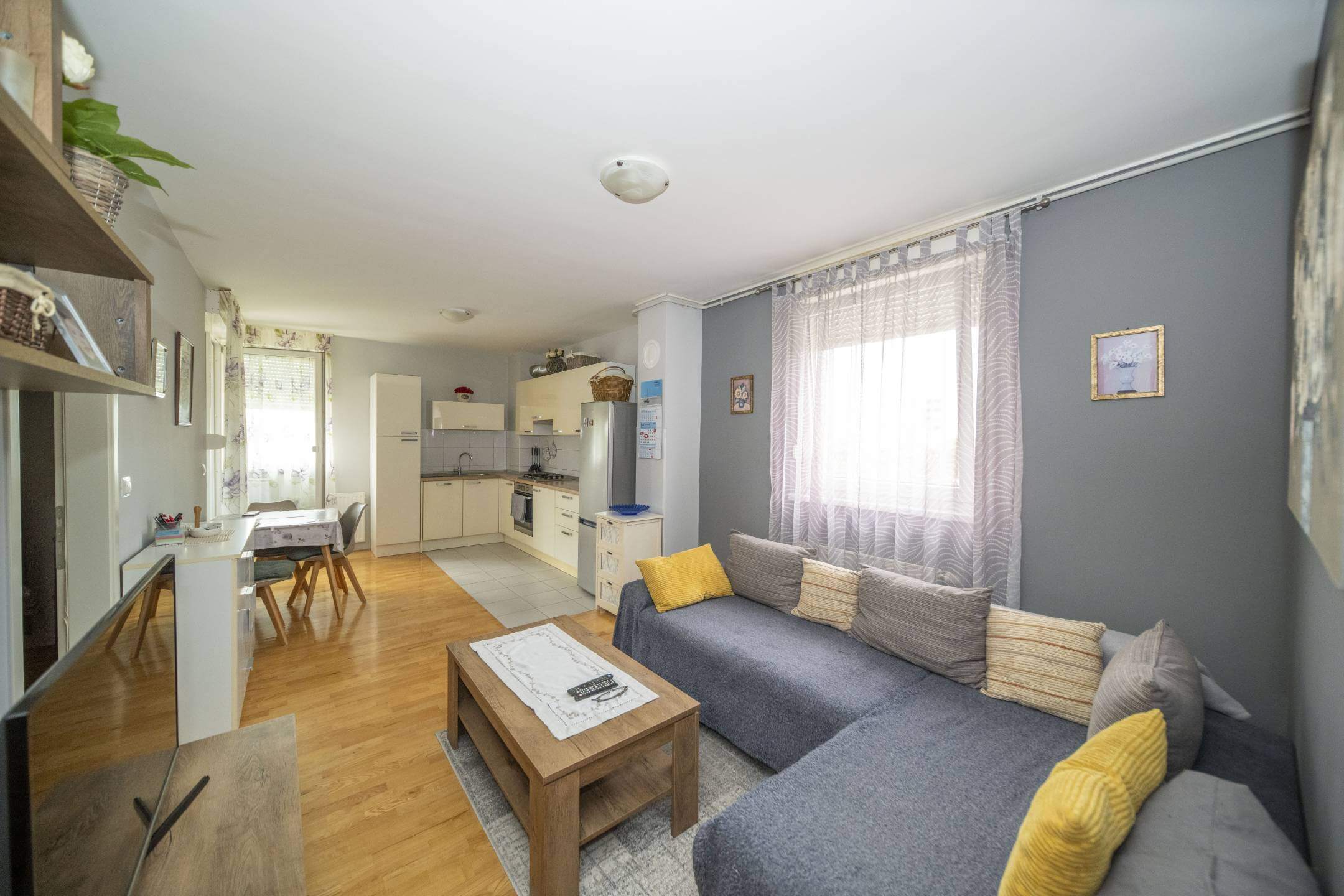 Otvoreni dnevni boravak sa sivom sofom u obliku slova L, žutim jastucima i drvenim stolićem za kavu; kuhinja u pozadini.