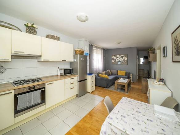 Otvorena kuhinja i dnevni boravak: krem ormarići, hladnjak od nehrđajućeg čelika i siva sofa sa žutim jastucima u pozadini.