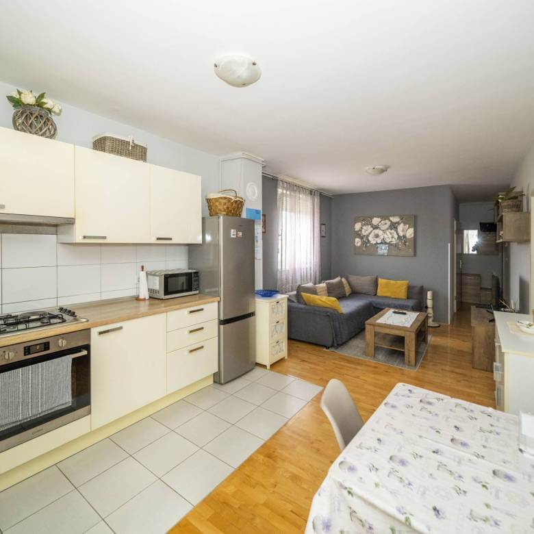 Otvorena kuhinja i dnevni boravak: krem ormarići, hladnjak od nehrđajućeg čelika i siva sofa sa žutim jastucima u pozadini.