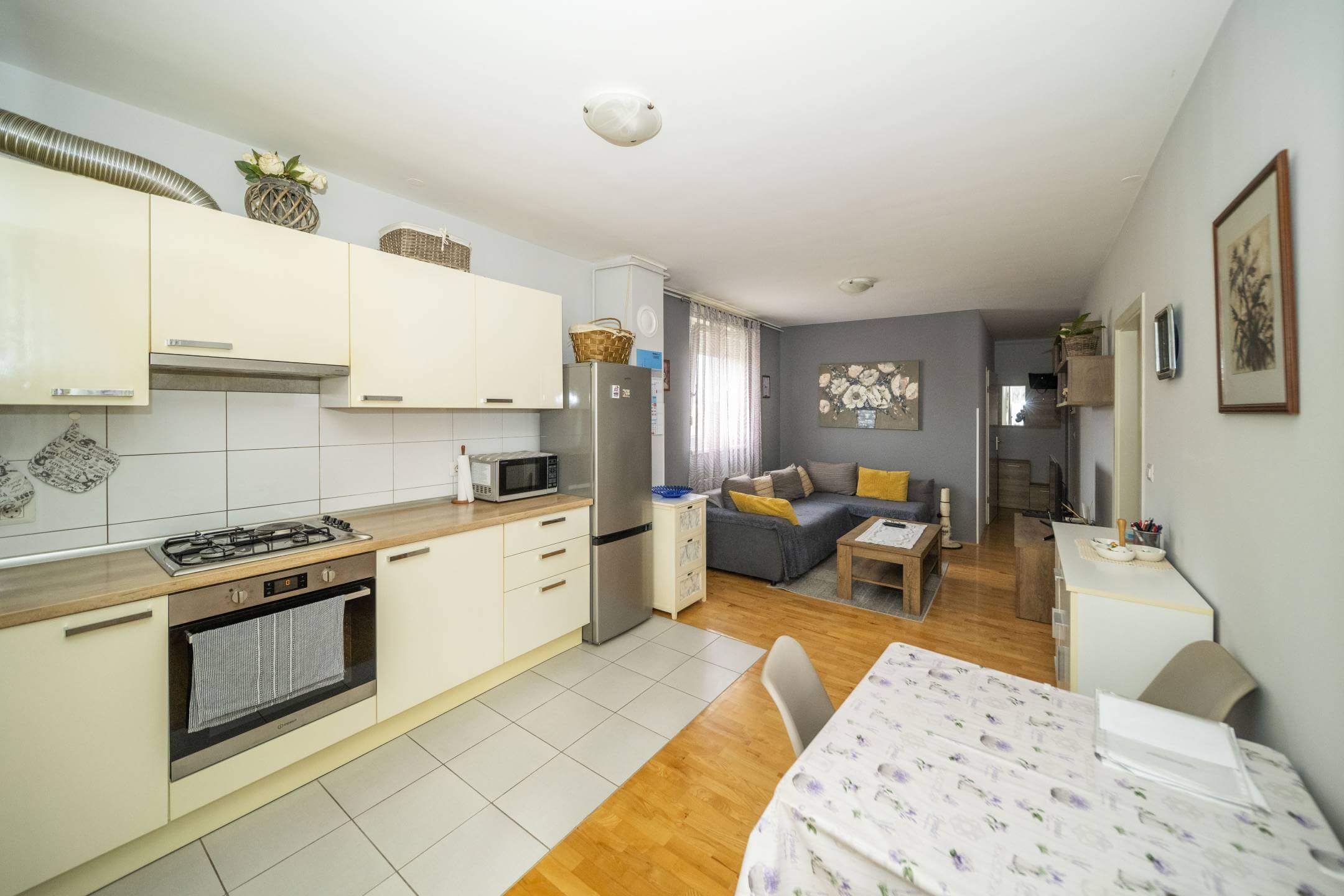 Otvorena kuhinja i dnevni boravak: krem ormarići, hladnjak od nehrđajućeg čelika i siva sofa sa žutim jastucima u pozadini.