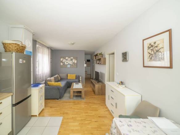 Otvoreni dnevni boravak i kuhinja sa sivim setom, žutim jastucima, drvenim stolićem za kavu i TV-om na drvenoj konzoli nasuprot sofe.