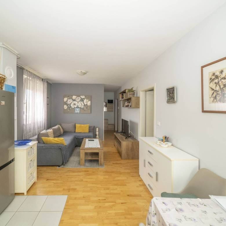 Otvoreni dnevni boravak i kuhinja sa sivim setom, žutim jastucima, drvenim stolićem za kavu i TV-om na drvenoj konzoli nasuprot sofe.