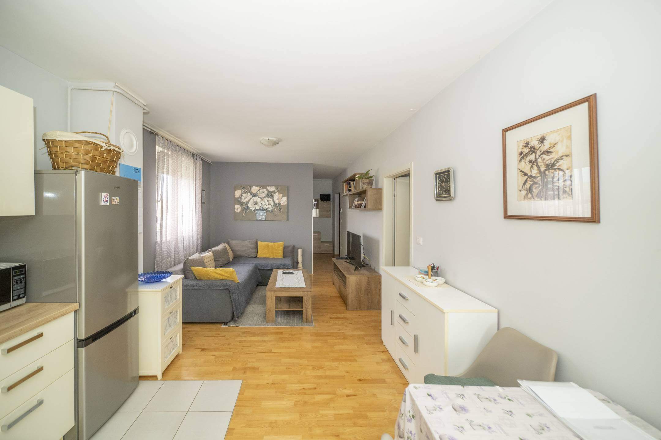 Otvoreni dnevni boravak i kuhinja sa sivim setom, žutim jastucima, drvenim stolićem za kavu i TV-om na drvenoj konzoli nasuprot sofe.