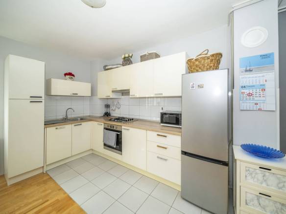 Svijetla kuhinja u obliku slova U s krem ormarićima, hladnjakom od nehrđajućeg čelika, sudoperom, plinskim štednjakom i kalendarom na zidu pored hladnjaka.