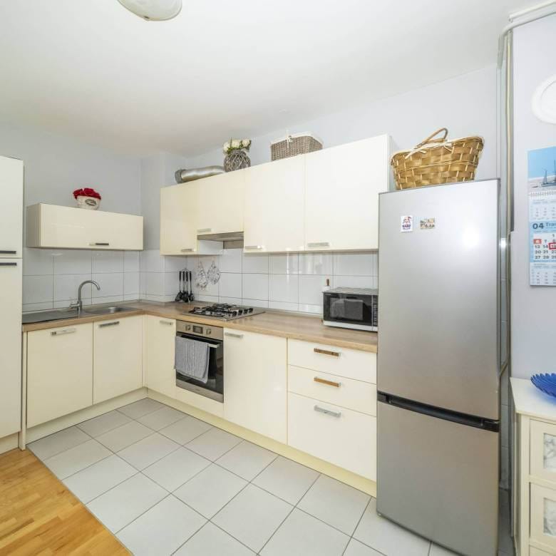 Svijetla kuhinja u obliku slova U s krem ormarićima, hladnjakom od nehrđajućeg čelika, sudoperom, plinskim štednjakom i kalendarom na zidu pored hladnjaka.