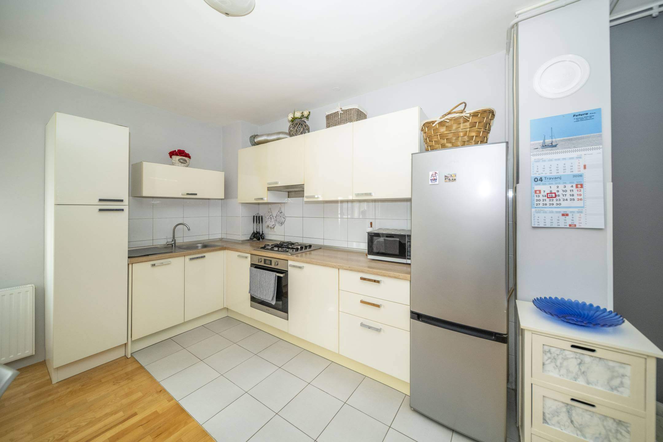 Svijetla kuhinja u obliku slova U s krem ormarićima, hladnjakom od nehrđajućeg čelika, sudoperom, plinskim štednjakom i kalendarom na zidu pored hladnjaka.