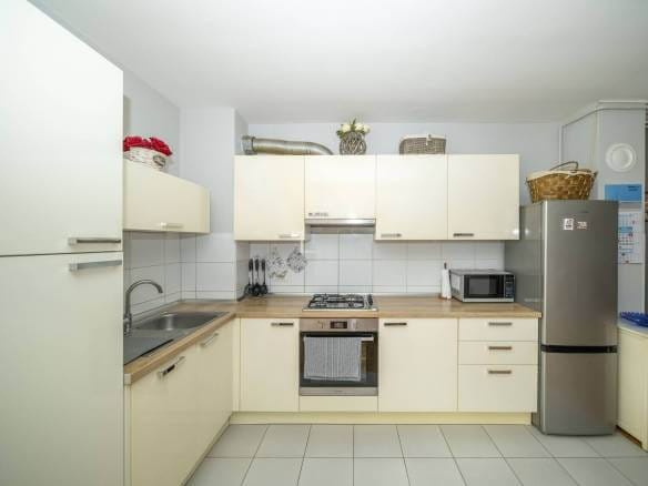 Moderna kuhinja u obliku slova L s krem ormarićima, drvenim radnim pločama i uređajima od nehrđajućeg čelika.