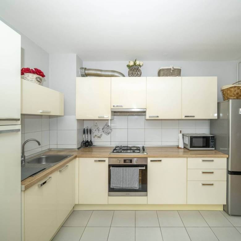 Moderna kuhinja u obliku slova L s krem ormarićima, drvenim radnim pločama i uređajima od nehrđajućeg čelika.