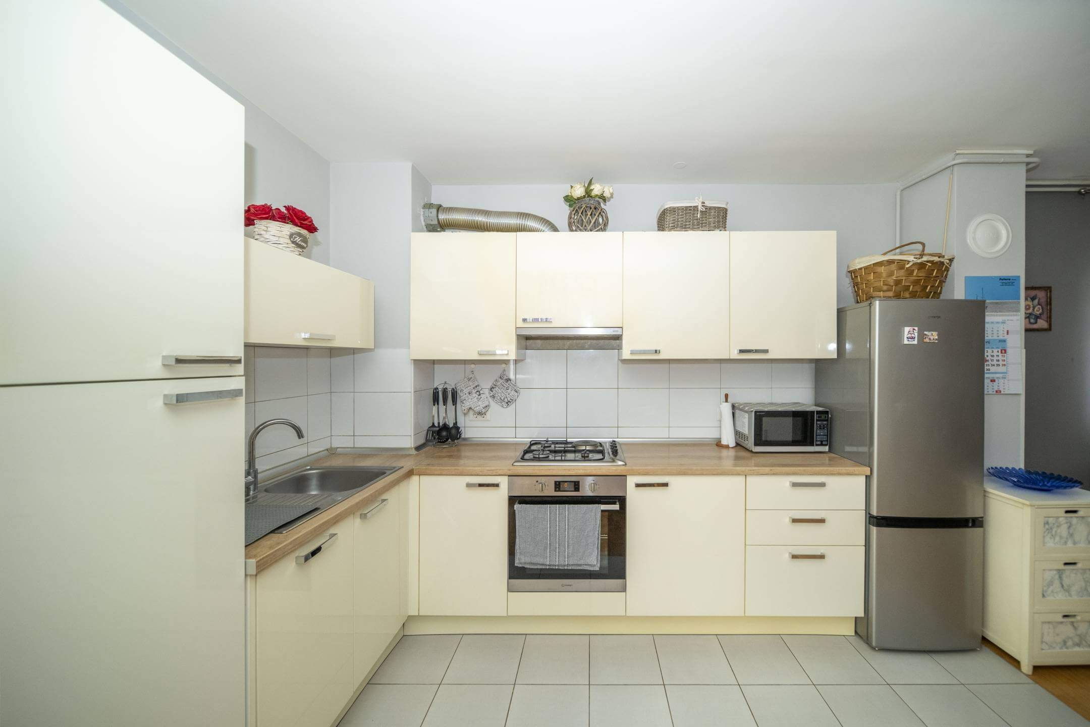 Moderna kuhinja u obliku slova L s krem ormarićima, drvenim radnim pločama i uređajima od nehrđajućeg čelika.