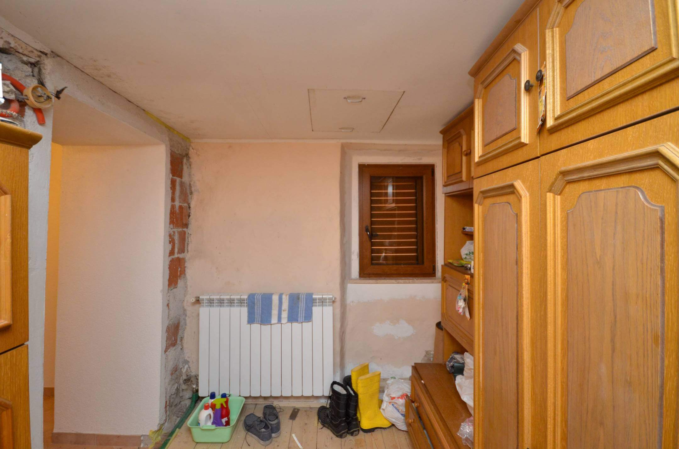 Ozka shramba za opremo z lesenimi omaricami na desni, radiatorjem, majhnim oknom in raztresenimi škornji ter čistilnimi sredstvi po tleh.