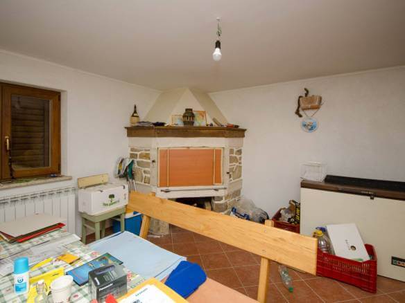 Natrpana domača delavnica s kamnitim kaminom, lesenim tramom v ospredju in mizo, prekrito s papirji, steklenicami in orodjem pod eno samo visečo žarnico.