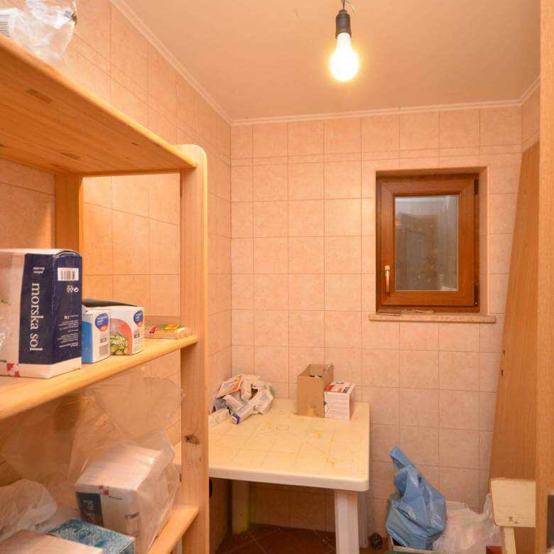 Majhna shramba z lesenimi policami, raztresenimi škatlami, plastičnimi vrečkami in golo žarnico, ki visi s stropa.