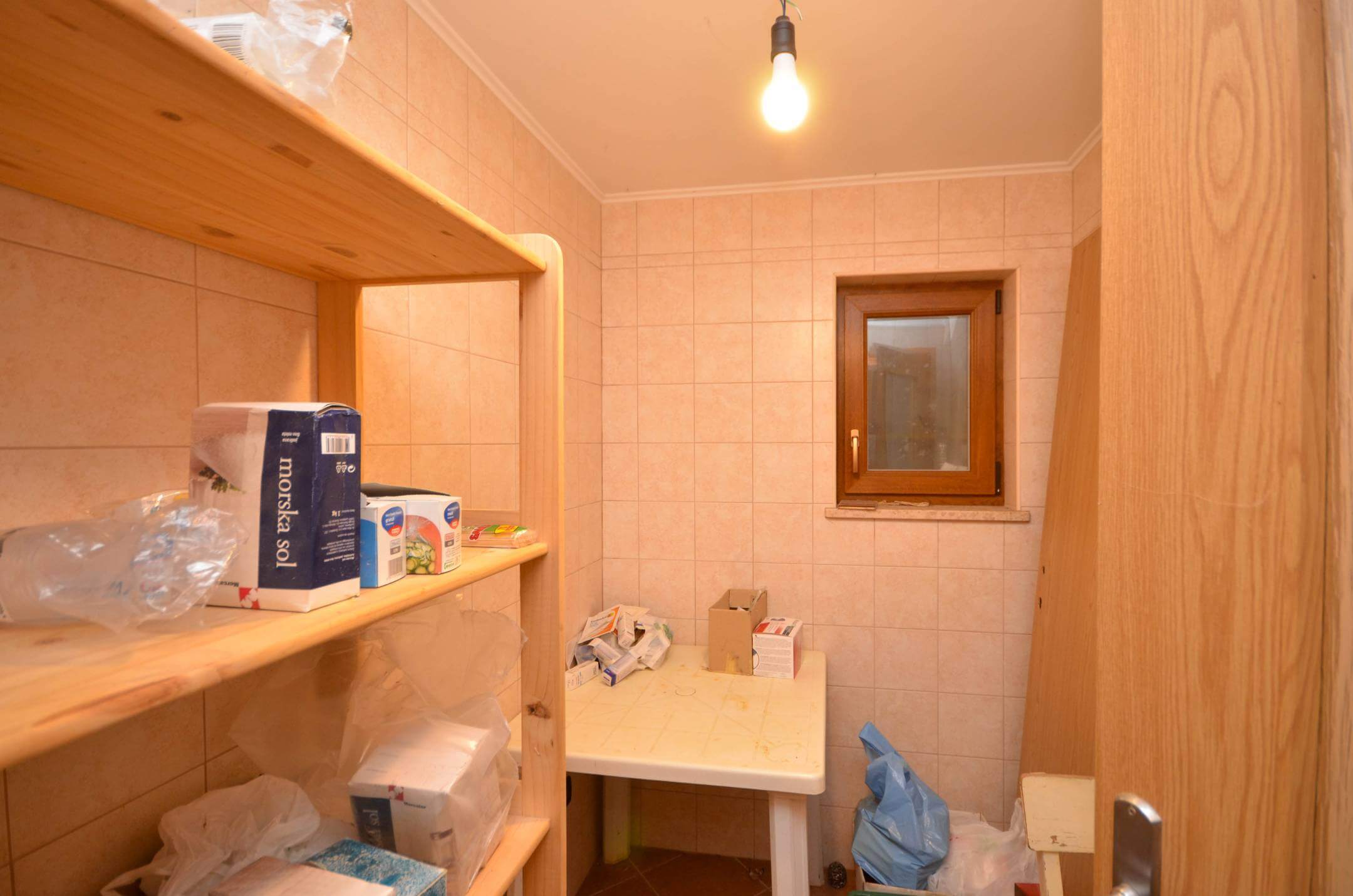 Majhna shramba z lesenimi policami, raztresenimi škatlami, plastičnimi vrečkami in golo žarnico, ki visi s stropa.