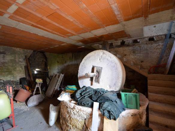 Prašna kletna delavnica z velikim okroglim brusilnim kolesom, opečnatim stropom ter raztresenim orodjem in cevmi.